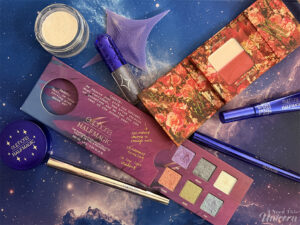 Flatlay of the Half Magic Beauty x Euphoria makeup collection featuring glitter eyeshadows, blue eyeliner, highlighter balm, and editorial makeup packaging on a galaxy background.
