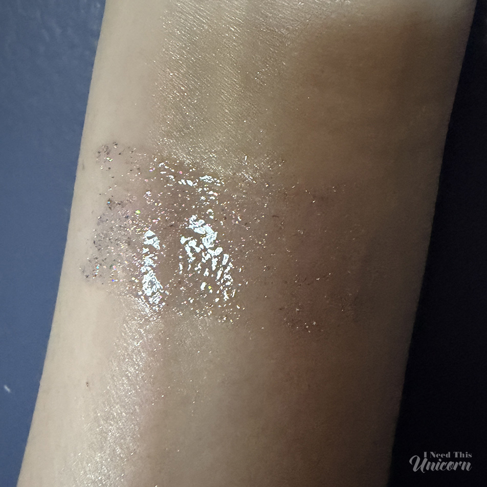 Digital Doll holographic lip gloss by Queen Cosmetics showing a sheer holographic sparkle swatch on skin.