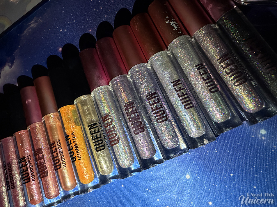 Queen Cosmetics Tester Diamond Shimmer Glosses | I Need This Unicorn Large Queen Cosmetics lip gloss collection arranged in a row on a galaxy-themed background. The collection includes holographic and multichrome glosses in white, pink, lavender, gold, bronze, and black-based shades. Several well-loved tubes show wear from frequent use and are included as comparison references for the new tester shades.