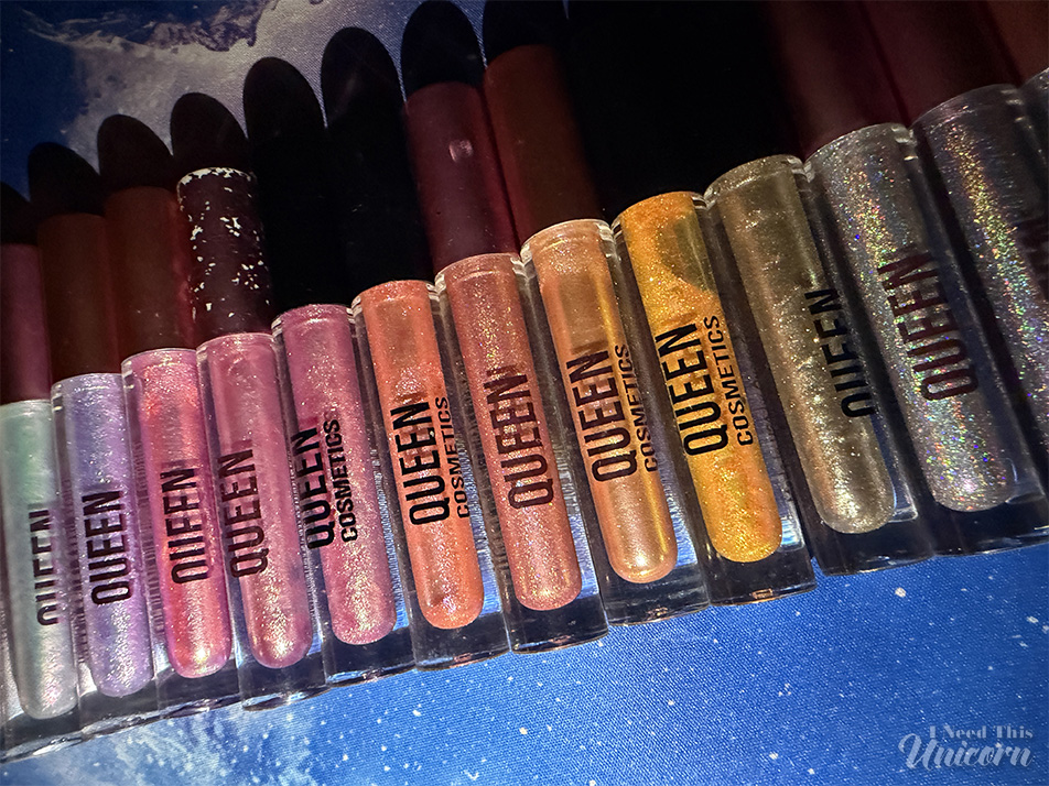 Queen Cosmetics Tester Diamond Shimmer Glosses | I Need This Unicorn Queen Cosmetics lip gloss collection displayed on a galaxy desk mat. The lineup includes holographic, iridescent, glitter, and multichrome shades collected over several years and serves as a reference for comparing the new limited-edition tester glosses.