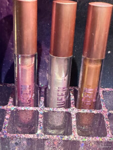Queen Cosmetics tester lip glosses photographed on a galaxy-themed background and inside a purple resin glitter lipstick holder highlighting holographic sparkle and multichrome shifts.