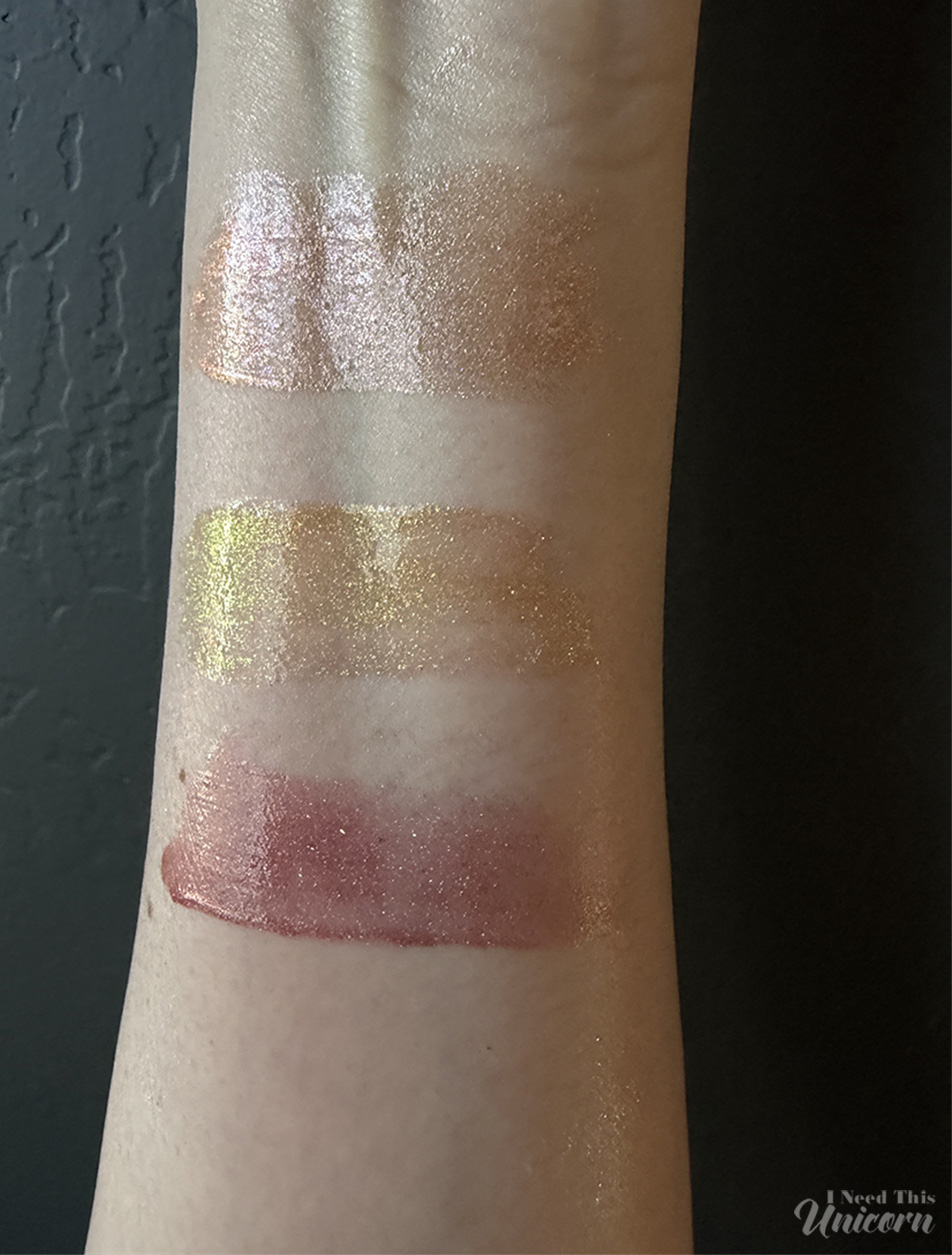 Queen Cosmetics Tester Diamond Shimmer Glosses | I Need This Unicorn Indoor lighting comparison of Queen Cosmetics Tester #3, Aphrodite, and Face Time swatched on the arm. Tester #3 reflects a soft champagne-pink glow, Aphrodite leans warm golden bronze, and Face Time shows a sheer rosy nude base with subtle sparkle.
