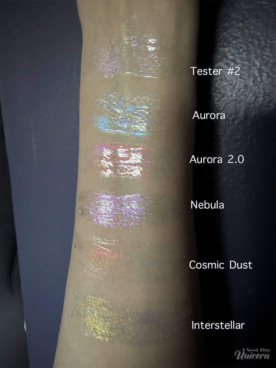 Queen Cosmetics Tester Diamond Shimmer Glosses | I Need This Unicorn Queen Cosmetics Tester #2 compared with Stardom Collection glosses in low indoor lighting. Swatches on bare skin show Tester #2's soft pink-lilac shimmer alongside Aurora's icy blue reflect, Aurora 2.0's peachy-pink glow, Nebula's lavender sparkle, Cosmic Dust's coral-orange shift, and Interstellar's gold shimmer.