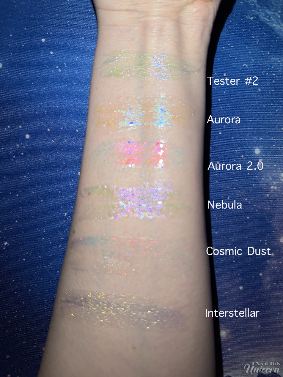 Queen Cosmetics Tester Diamond Shimmer Glosses | I Need This Unicorn Arm swatches of Queen Cosmetics Tester #2 and five Stardom Collection glosses photographed using direct smartphone flash in a dark room. The flash emphasizes reflective particles and color shifts, showing Tester #2's soft pink shimmer compared with Aurora's blue shift, Aurora 2.0's peach-pink shift, Nebula's lavender sparkle, Cosmic Dust's warm coral glow, and Interstellar's gold reflect.