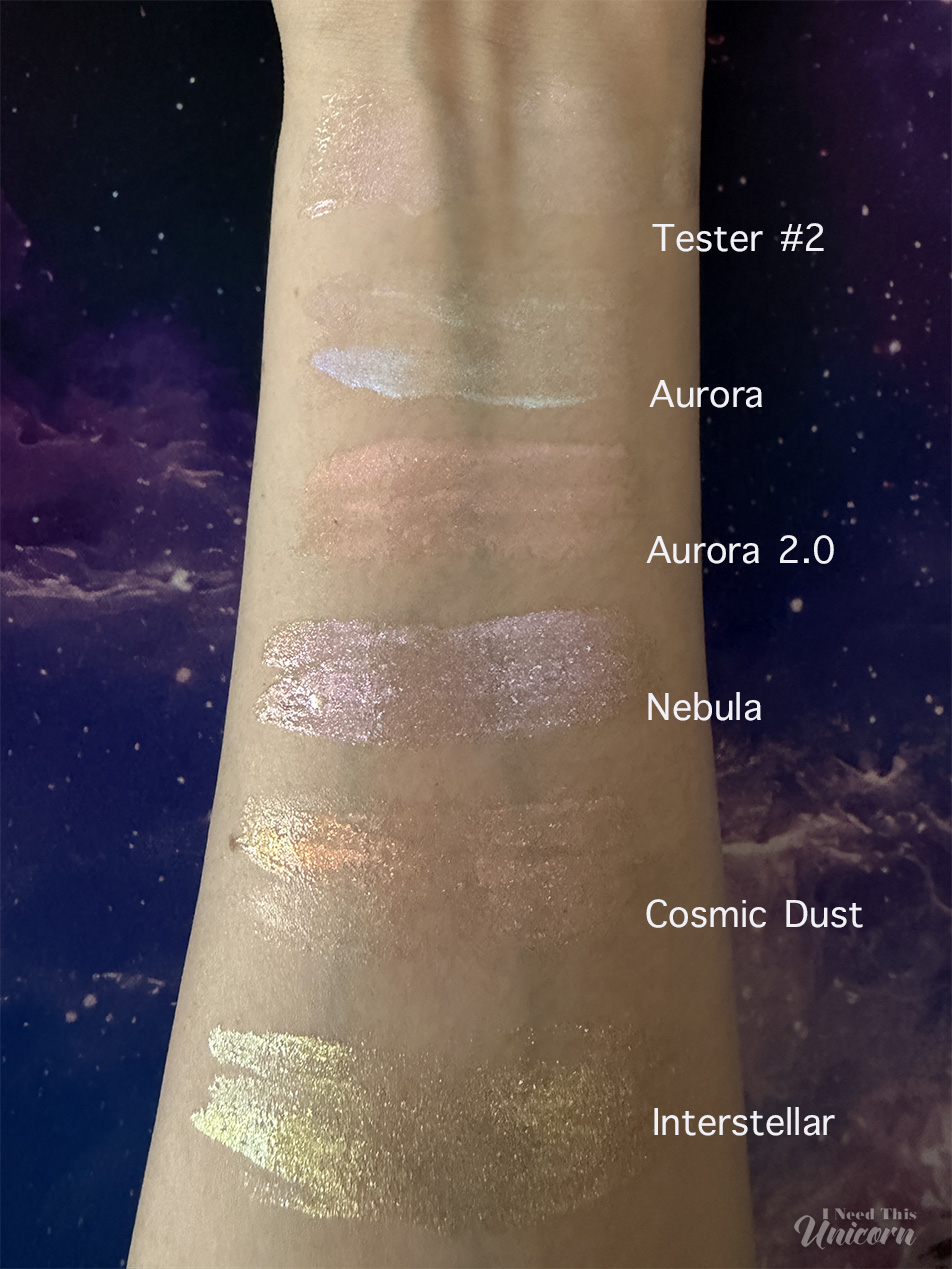 Queen Cosmetics Tester Diamond Shimmer Glosses | I Need This Unicorn Arm swatches of Queen Cosmetics Tester #2, Aurora, Aurora 2.0, Nebula, Cosmic Dust, and Interstellar photographed indoors. The lighting highlights each gloss's base color and reflective shimmer, showing Tester #2's pink-lavender pearl finish compared to the blue, peach, lavender, coral, and gold shifts of the Stardom Collection shades.